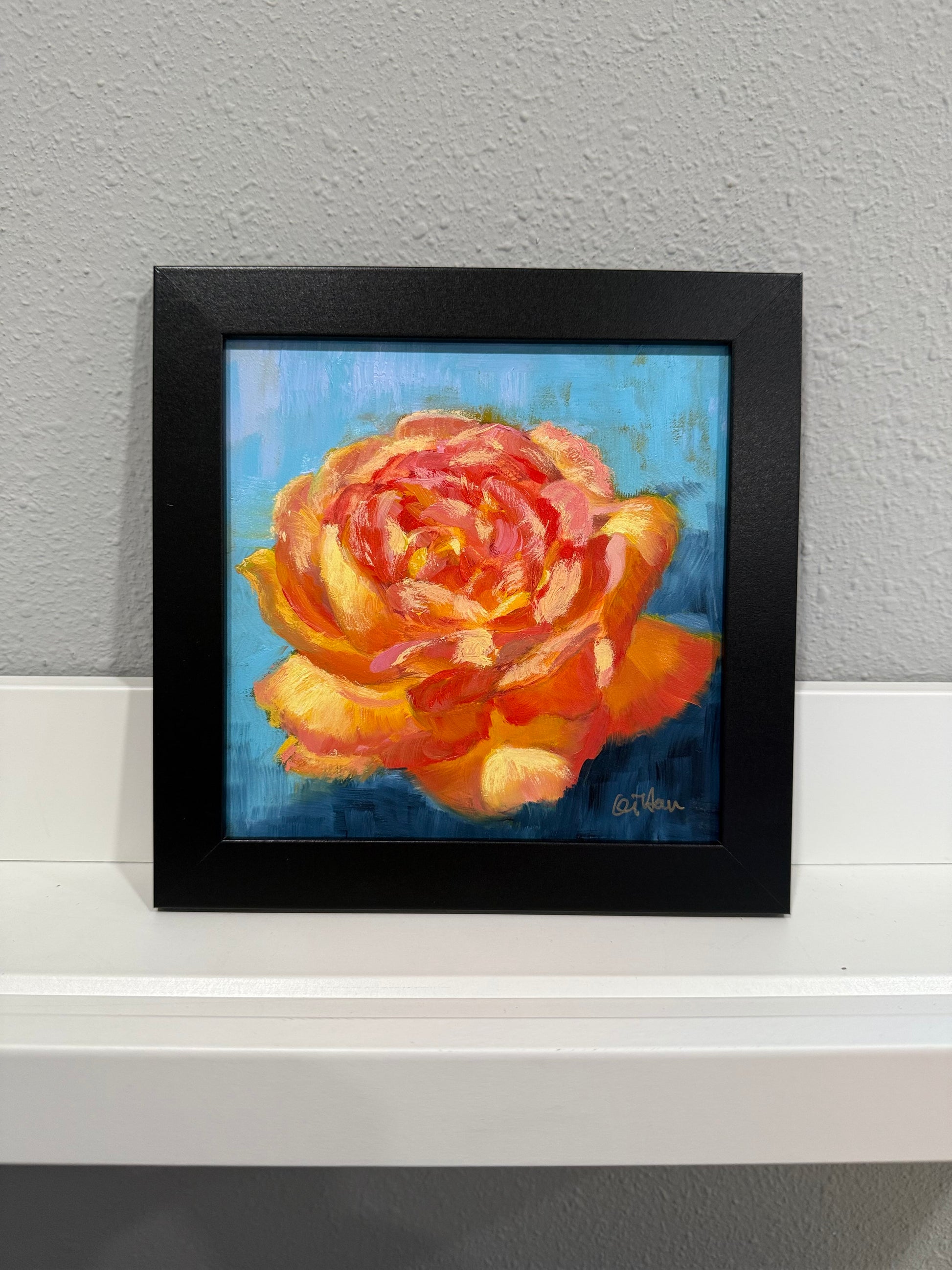 Framed painting of a rose on a white surface with a gray wall background