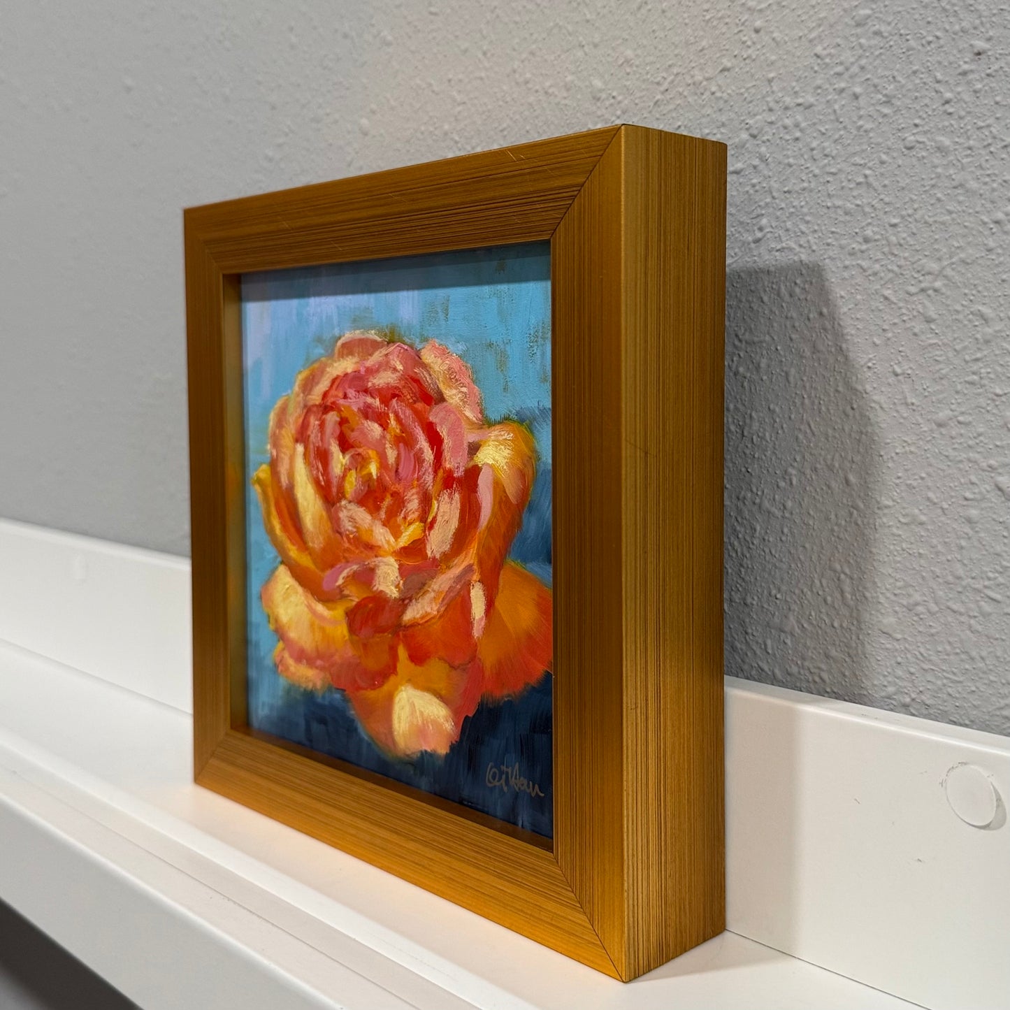 Framed painting of a rose on a white surface with a gray wall background