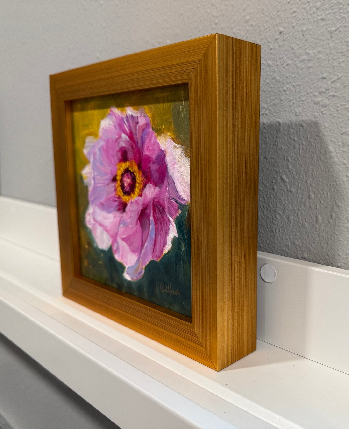 Framed painting of a pink flower on a white shelf against a gray wall