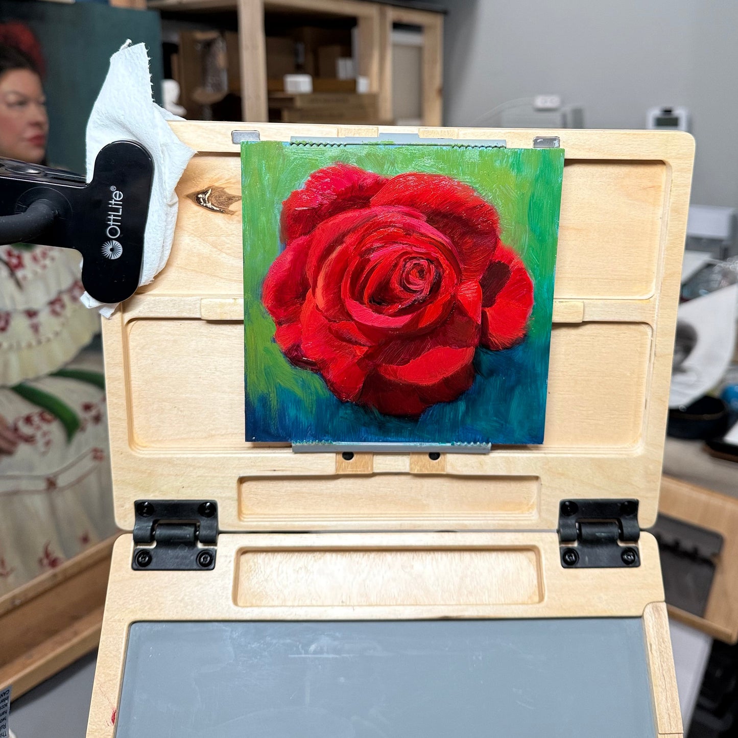 Small painting of a red rose on a wooden easel with a blurred background