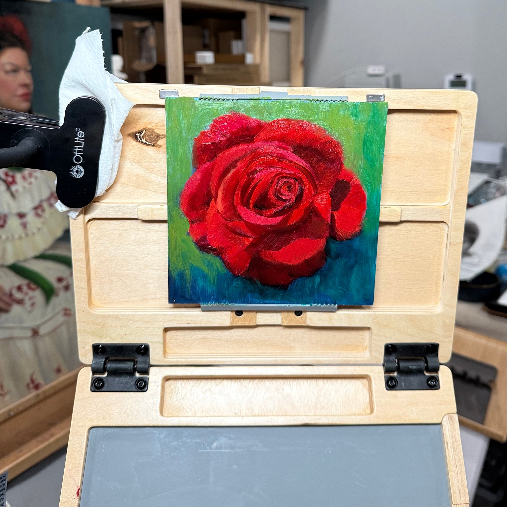 Small painting of a red rose on a wooden easel with a blurred background